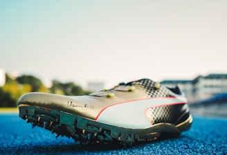 A track and field sports shoe is positioned on a blue running track, with soft sunlight illuminating its design. The shoe features sharp cleats underneath, a sleek metallic finish, and a signature inscribed on the side. Blurred buildings and greenery are visible in the background.