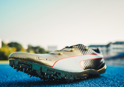 A track and field sports shoe is positioned on a blue running track, with soft sunlight illuminating its design. The shoe features sharp cleats underneath, a sleek metallic finish, and a signature inscribed on the side. Blurred buildings and greenery are visible in the background.