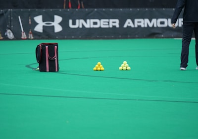 A sports field features a black and pink sports bag, two pyramids of yellow balls, and a portion of a person in dark athletic gear. The background displays a banner with the Under Armour logo.