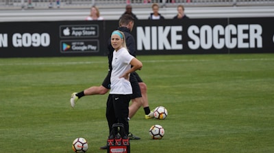 A person in sports attire stands on a soccer field, surrounded by several soccer balls and a carrier with bottles. The field is bordered by advertisements for Nike Soccer and app stores. Several people sit in bleachers in the background.