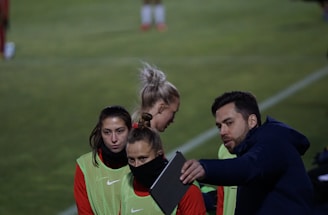A group of individuals on a sports field are wearing green vests over red attire. One person is holding a tablet and seems to be discussing or showing something to the others. The atmosphere suggests a sports event, likely soccer, indicated by their clothing and the setting. The background is a grassy field.