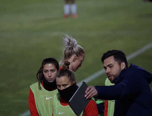 A group of individuals on a sports field are wearing green vests over red attire. One person is holding a tablet and seems to be discussing or showing something to the others. The atmosphere suggests a sports event, likely soccer, indicated by their clothing and the setting. The background is a grassy field.