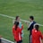 A group of people wearing sports attire are gathered on a grassy field. Some individuals are dressed in red jackets, while others are in turquoise and navy blue. A coach appears to be giving instructions. Sports equipment, such as water bottles and a bench with towels, can be seen nearby.