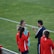 A group of people wearing sports attire are gathered on a grassy field. Some individuals are dressed in red jackets, while others are in turquoise and navy blue. A coach appears to be giving instructions. Sports equipment, such as water bottles and a bench with towels, can be seen nearby.