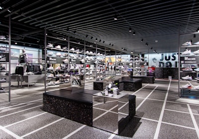 A modern sneaker store with sleek black shelving displaying various athletic shoes and sports apparel. Central seating areas and spotlights provide a contemporary retail ambiance. The signage prominently features a famous sportswear slogan in the background.