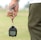 A person is holding a stopwatch in their left hand with a strap wrapped around their wrist. The background is an out-of-focus field or grass area. The person is wearing a white shirt and dark green sports pants.