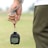 A person is holding a stopwatch in their left hand with a strap wrapped around their wrist. The background is an out-of-focus field or grass area. The person is wearing a white shirt and dark green sports pants.
