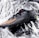 A black sports shoe with orange stripes and a sleek design lies on a crumpled, reflective silver surface. The shoe features textured fabric and a small green accent at the heel.