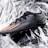 A black sports shoe with orange stripes and a sleek design lies on a crumpled, reflective silver surface. The shoe features textured fabric and a small green accent at the heel.